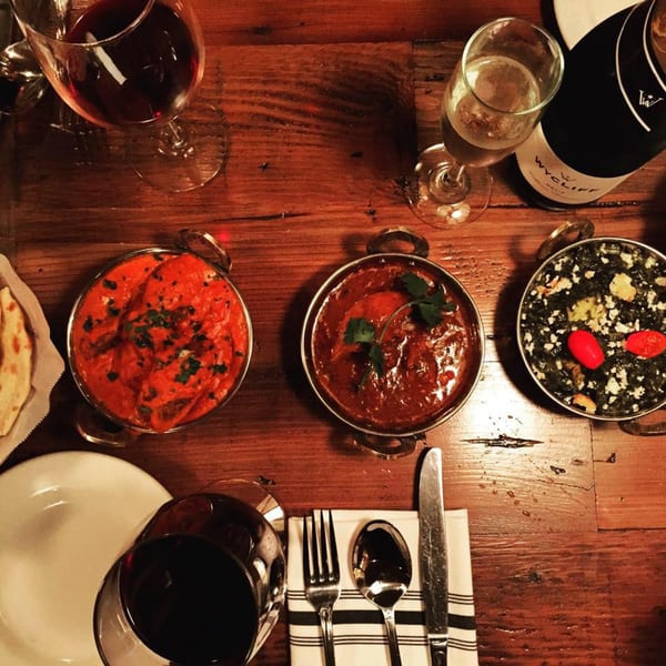 Overhead view of three Indian dishes, naan, wine, and cutlery on a rustic wooden table.